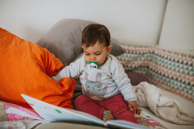 Cute baby boy reading book while sitting on bed at home