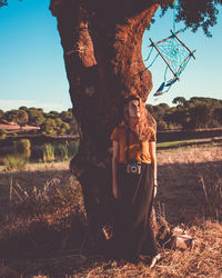 Woman standing by tree trunk on field against sky