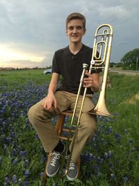 Portrait of happy young man holding saxophone while sitting on stool amidst flowers against cloudy sky