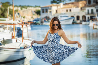 Young brunette woman standing on a dock of cala figuera harbour, wearing a blue floral pattern dress