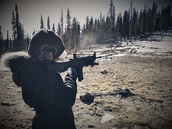 Woman aiming rifle while standing in forest during winter