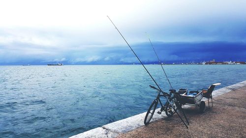 Fishing rods with bicycle and chair on promenade by sea against cloudy sky