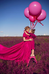 Mother and daughter in pink dresses are standing on a field with flowers at sunset in summer