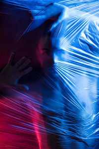 Pensive young handsome shirtless muscular male looking at camera while putting hands on surface of big transparent tape in light of blue and red lamps