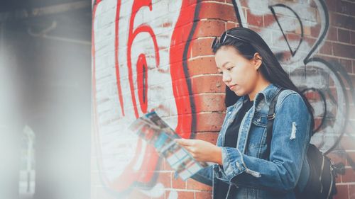 Side view of woman reading magazine while standing against graffiti wall