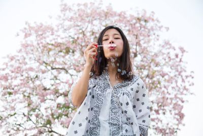 Portrait of beautiful woman blowing bubbles while standing against cherry blossom tree
