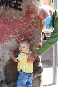 Cute boy standing with grandfather by wall outdoors