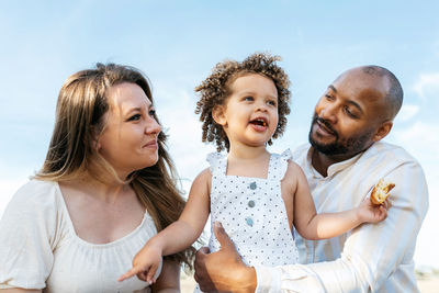 Low angle of happy multiethnic family with cute little daughter enjoying summer picnic in nature