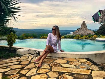 Full length of woman sitting on retaining wall by swimming pool against sky