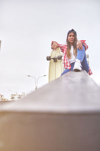 Beautiful young woman in cap is sitting at skatepark on the ramp with her longboard.