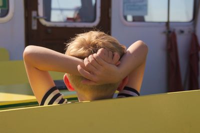 Rear view of boy with hands behind head sitting on bench against bus