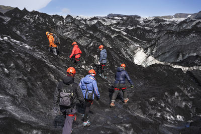 Back view group of unrecognizable travelers with warm clothes and protective helmets walking on glacier covered with volcanic ash during trekking in iceland