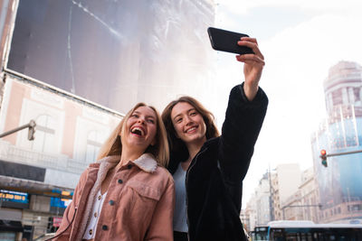Low angle of cheerful female friends standing on street in madrid and taking self shot on smartphone during city stroll