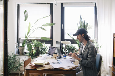 Side view of male architect using smart phone while sitting at desk in home office