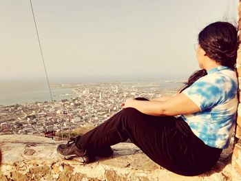 Rear view of woman sitting on cityscape against sky