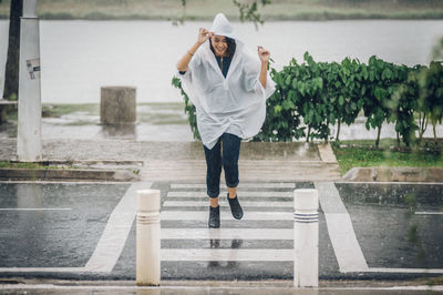 Smiling woman wearing raincoat crossing street against lake during monsoon