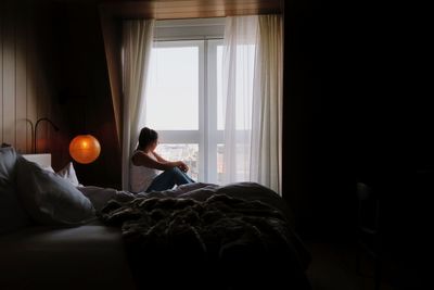 Side view of woman sitting on window in bedroom at home