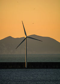 Silhouette wind turbine by sea against orange sky