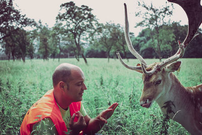 Side view of mature man crouching by deer on grassy field in forest