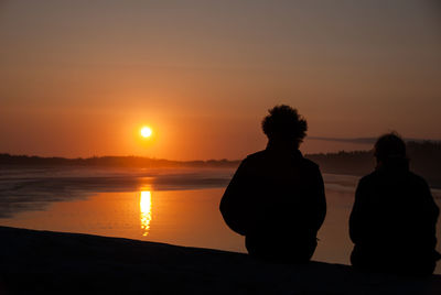 Silhouette couple sitting on shore against orange sunset sky