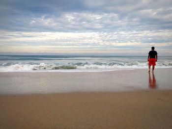 Rear view of man standing at beach against cloudy sky