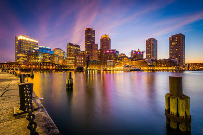 Illuminated modern buildings by river against sky at dusk