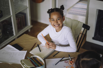 High angle portrait of happy girl sitting at table holding colored pencil