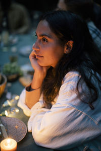 Woman with hand on chin sitting at dining table by illuminated candle