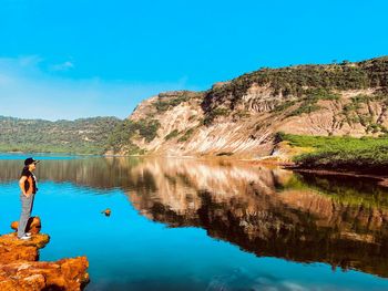 Woman standing by lake on rock against blue sky