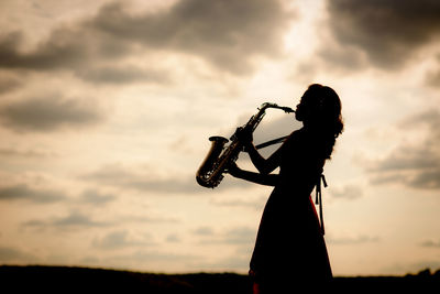 Side view of woman playing saxophone against cloudy sky during sunset