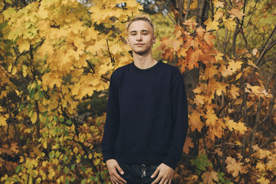 Confident teenage boy with blond hair standing against maple trees in autumn