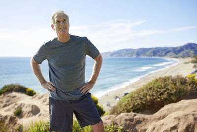 Smiling senior man with hands on hips standing on cliff at beach against sky