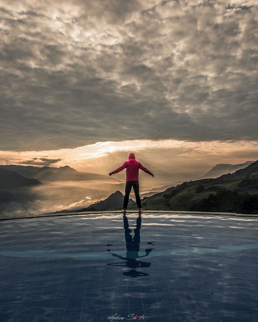 REAR VIEW OF MAN STANDING ON MOUNTAIN AGAINST SKY