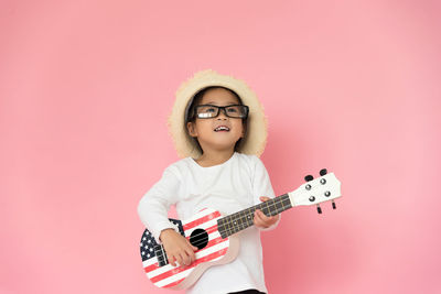 Girl wearing eyeglasses and hat while playing guitar against pink background