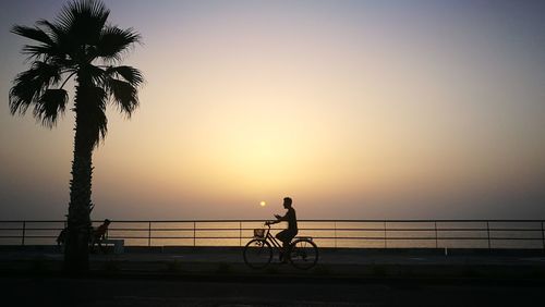 Silhouette people riding bicycle on sea against sky during sunset