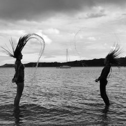 Mother and daughter tossing hair while standing on shore at beach against cloudy sky