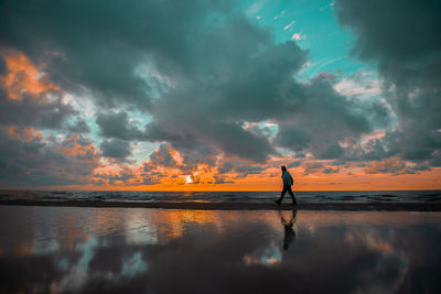 Man walking at beach against cloudy sky during sunset