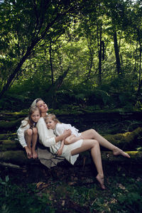 Family blonde mother with two children in a white dress is lying on tree log in the forest barefoot