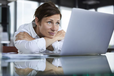 Businesswoman leaning on glass table in office looking at laptop