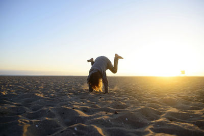 Girl practicing handstand at beach against clear sky during sunrise