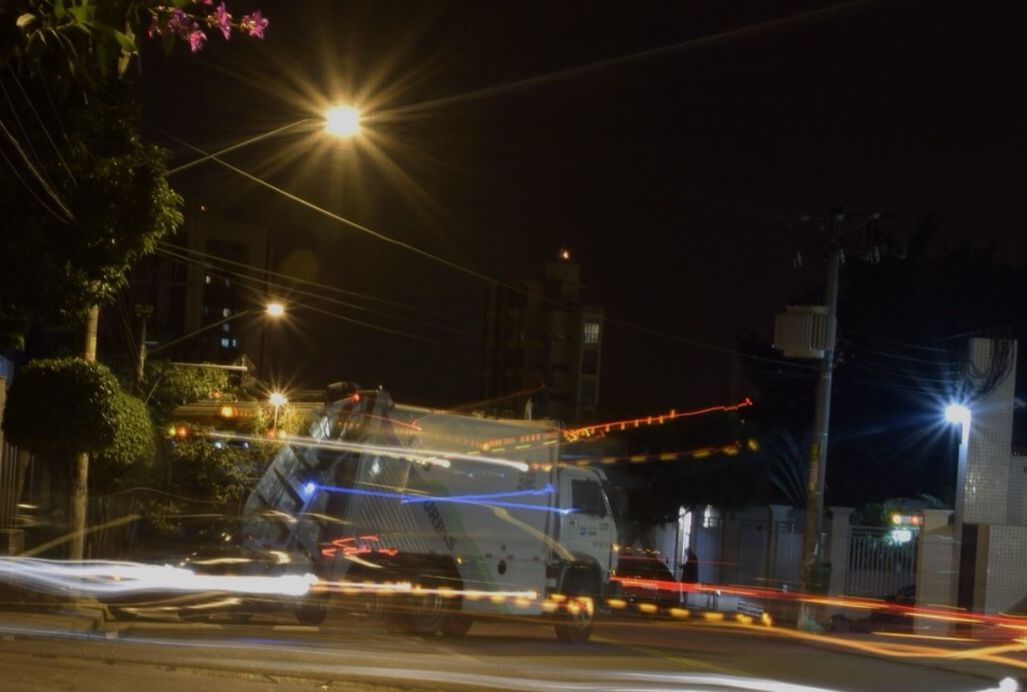 illuminated, night, street light, transportation, lighting equipment, mode of transport, street, land vehicle, car, road, sky, outdoors, light - natural phenomenon, built structure, building exterior, light beam, incidental people, tree, city, electric light