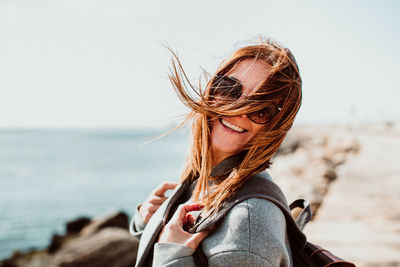 Side view portrait of cheerful woman with tousled hair standing on pier over sea against clear sky