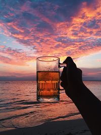 Woman hand holding beer glass against sea during sunset