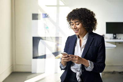 Smiling businesswoman using smart phone while standing against wall in office