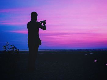 Silhouette man photographing sea while standing at beach against sky during sunset