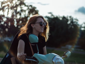 Young woman wearing sunglasses sitting on motor scooter against trees