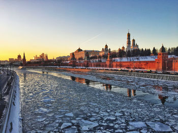 Frozen river by buildings against sky during sunset in city