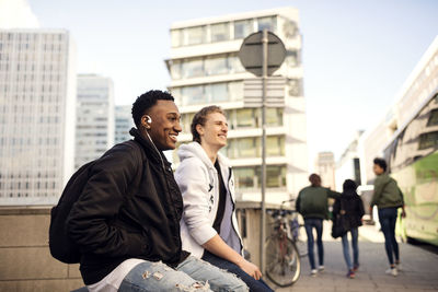 Happy teenagers resting on railing while friends walking on footpath in city