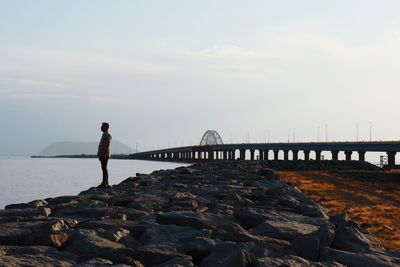 Man standing on rock by bridge against sky