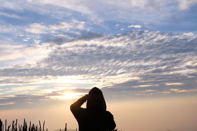 Silhouette person shielding eyes while standing against sky during sunset